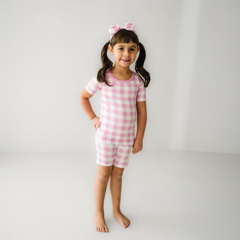 Click to see full screen - Alternate image of a child wearing a Pink Gingham two piece short sleeve and shorts pajama set paired with a matching luxe bow headband