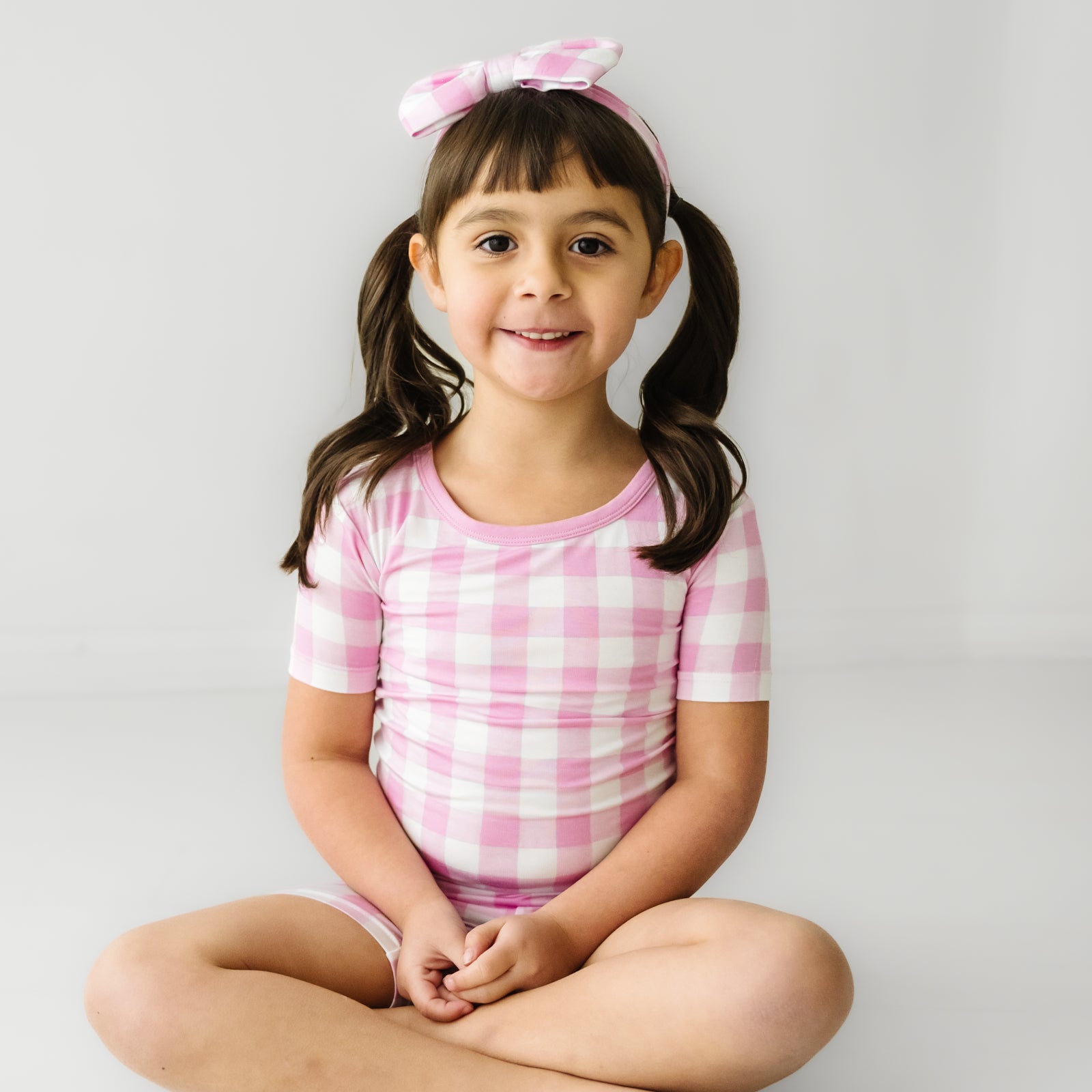 Child sitting wearing a Pink Gingham two piece short sleeve and shorts pajama set paired with a matching luxe bow headband