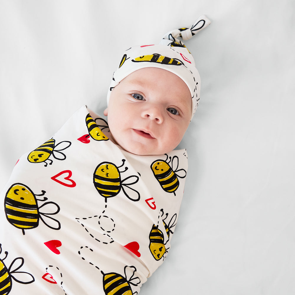 Close up image of a child swaddled in a Bee Mine swaddle and hat set on a bed