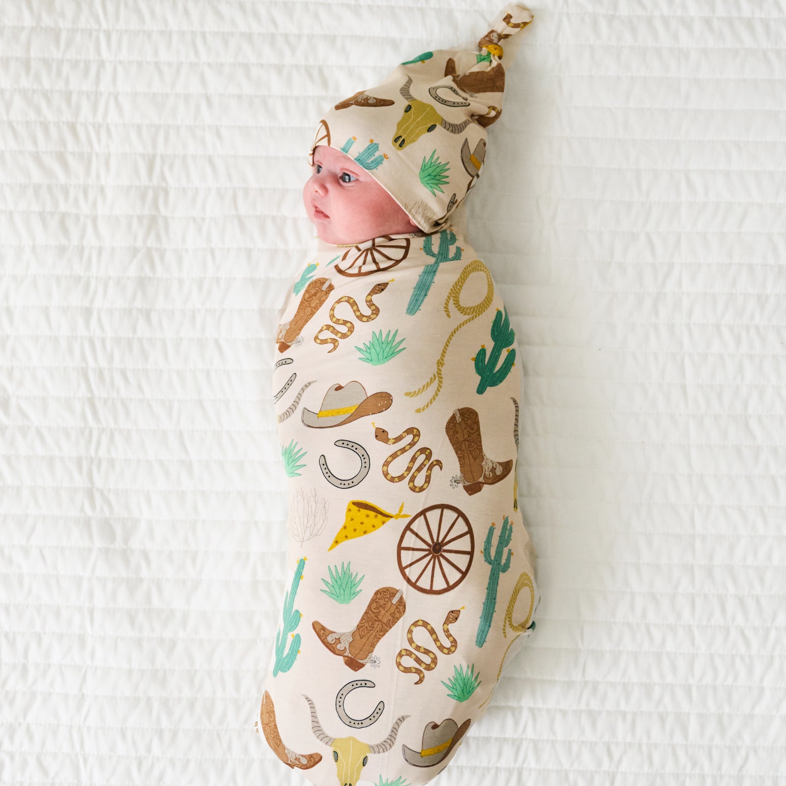 Child laying on a bed swaddled in a Caramel Ready to Rodeo swaddle and hat set