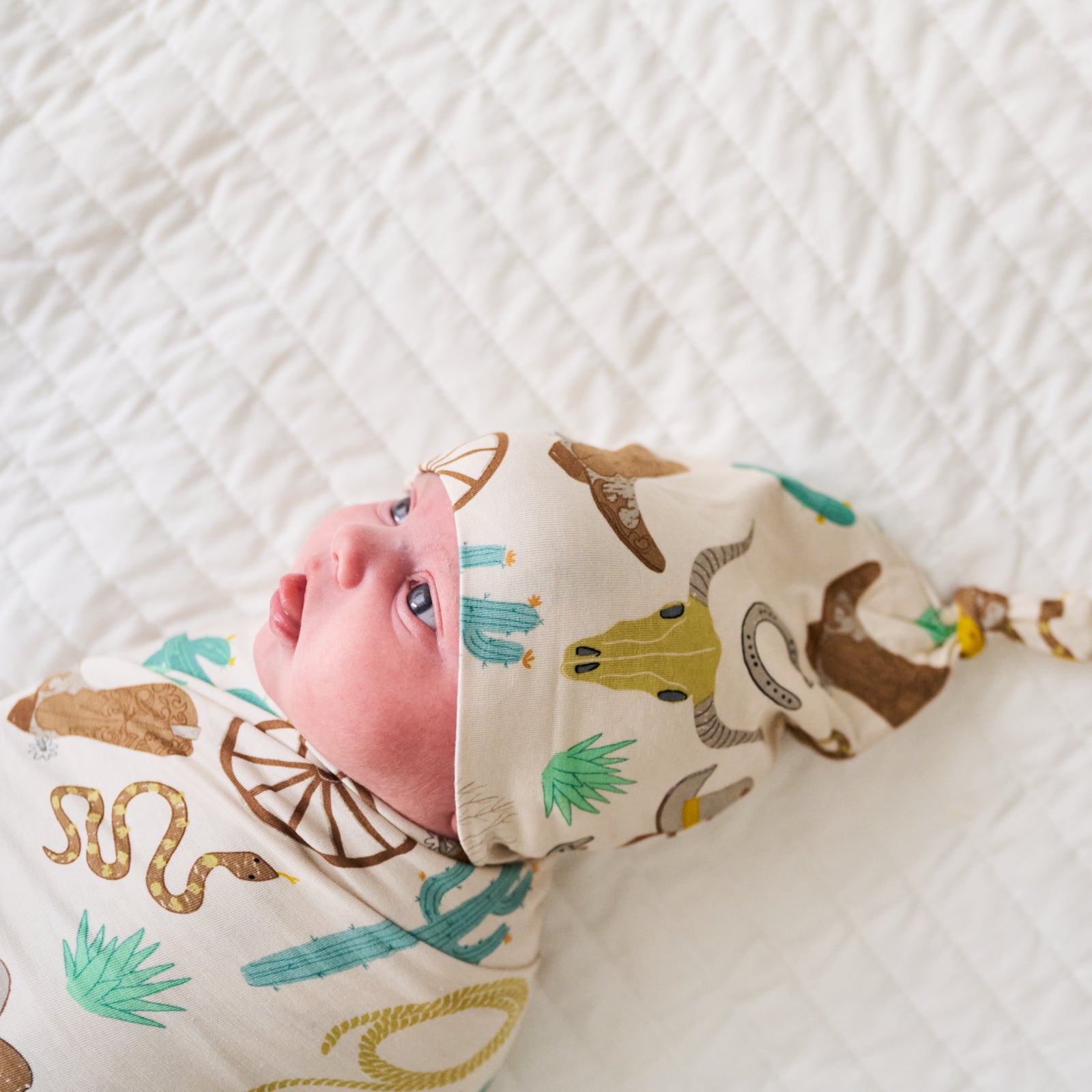 Close up image of a child laying on a bed swaddled in a Caramel Ready to Rodeo swaddle and hat set