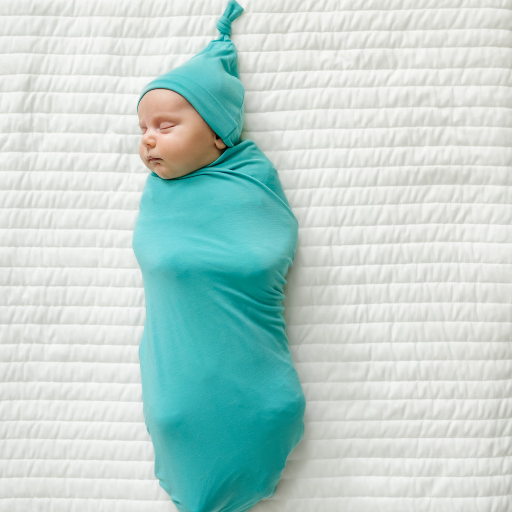 child swaddled on a bed wearing a Glacier Turquoise swaddle and hat set