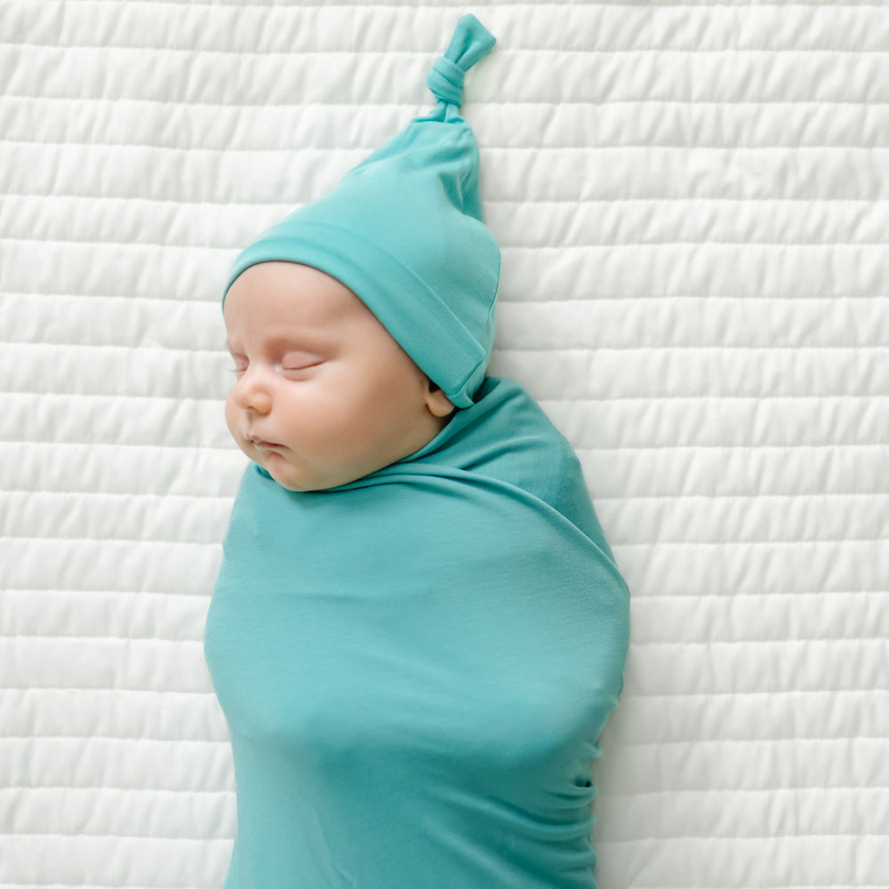 Close up image of a child swaddled on a bed wearing a Glacier Turquoise swaddle and hat set
