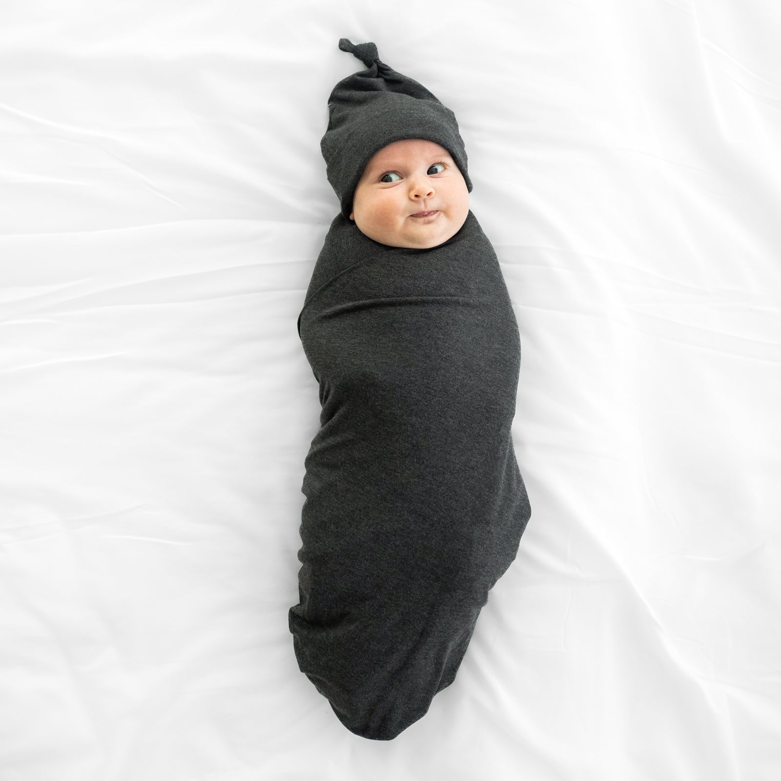 Child swaddled on a bed in a Cozy Heather Black swaddle and hat set