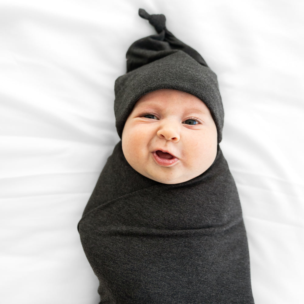 Close up image of a child swaddled on a bed in a Cozy Heather Black swaddle and hat set