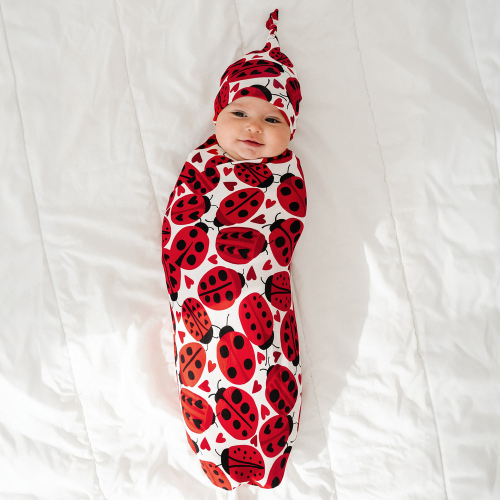 Click to see full screen - Child laying on a blanket swaddled in a Love Bug printed swaddle and hat set
