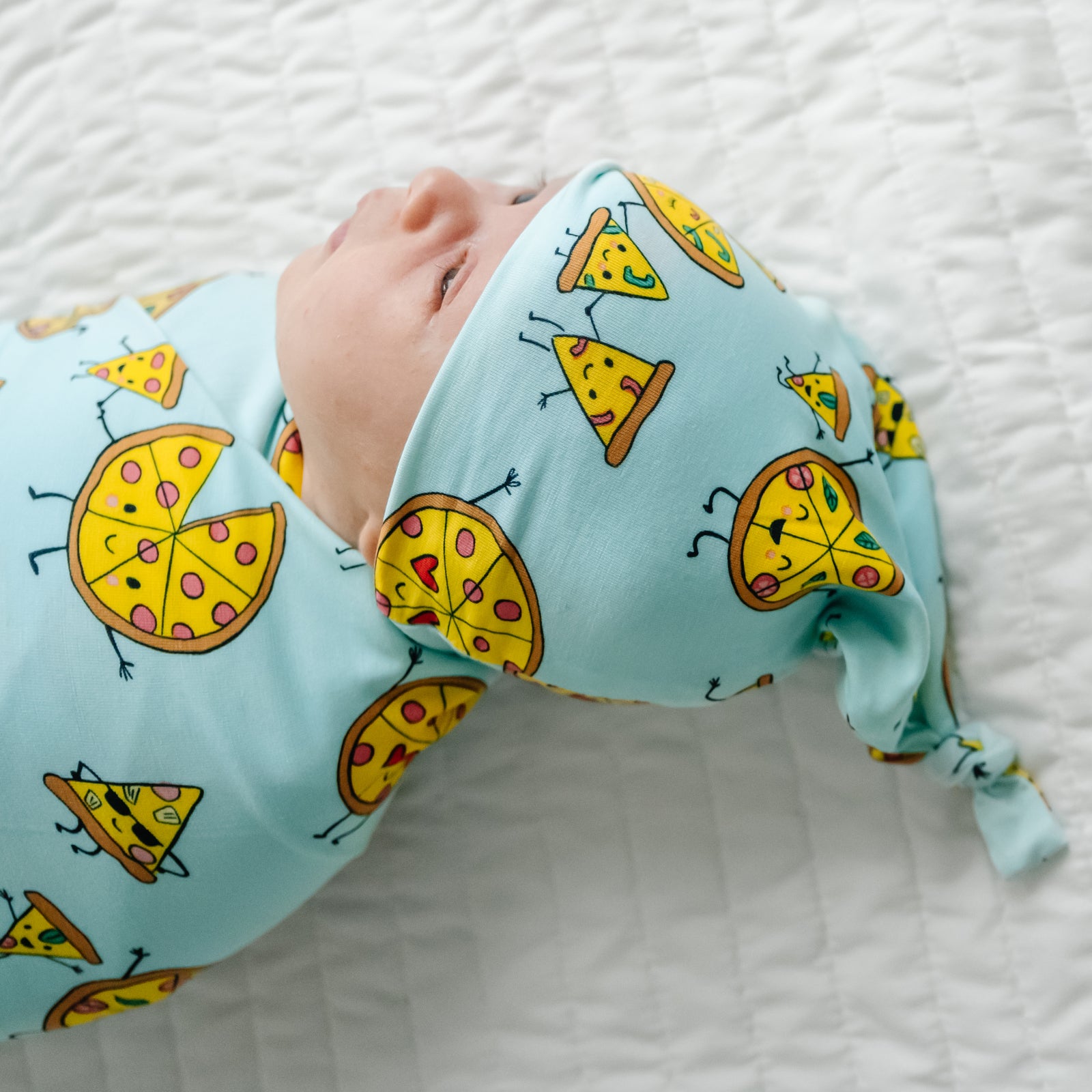Close up image of a child laying on a bed wearing a Pizza Pals swaddle and hat set