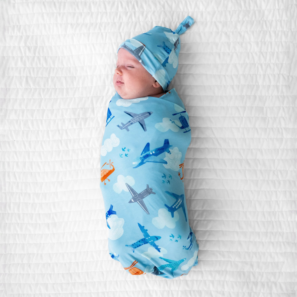 image of a child swaddled on a bed in a  Let's Fly swaddle and hat set