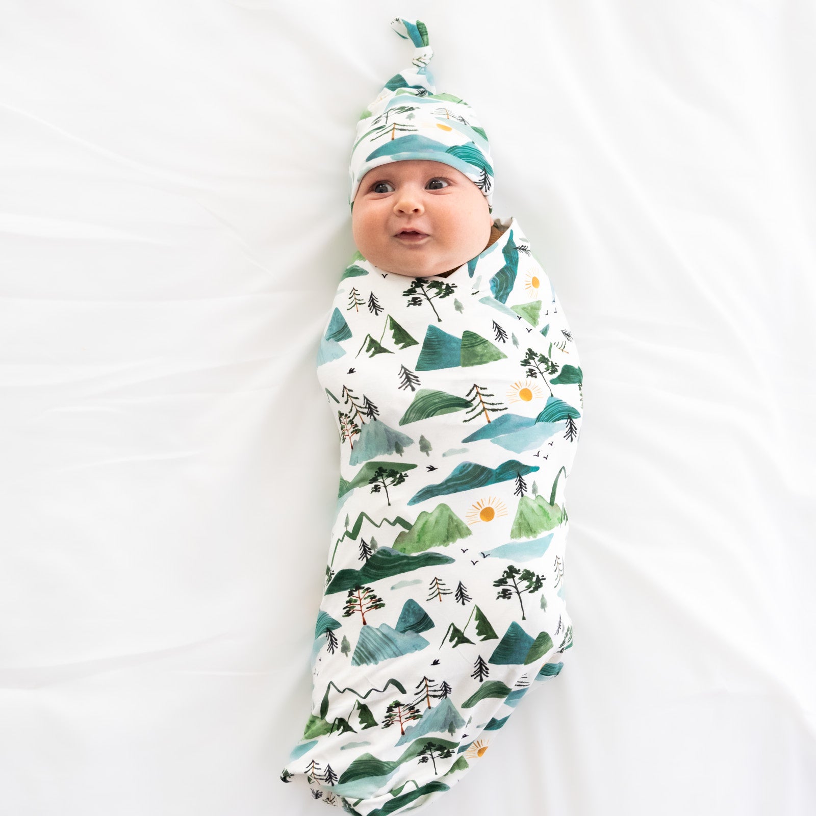 child laying on a bed swaddled in a Mountain Mist swaddle and hat set