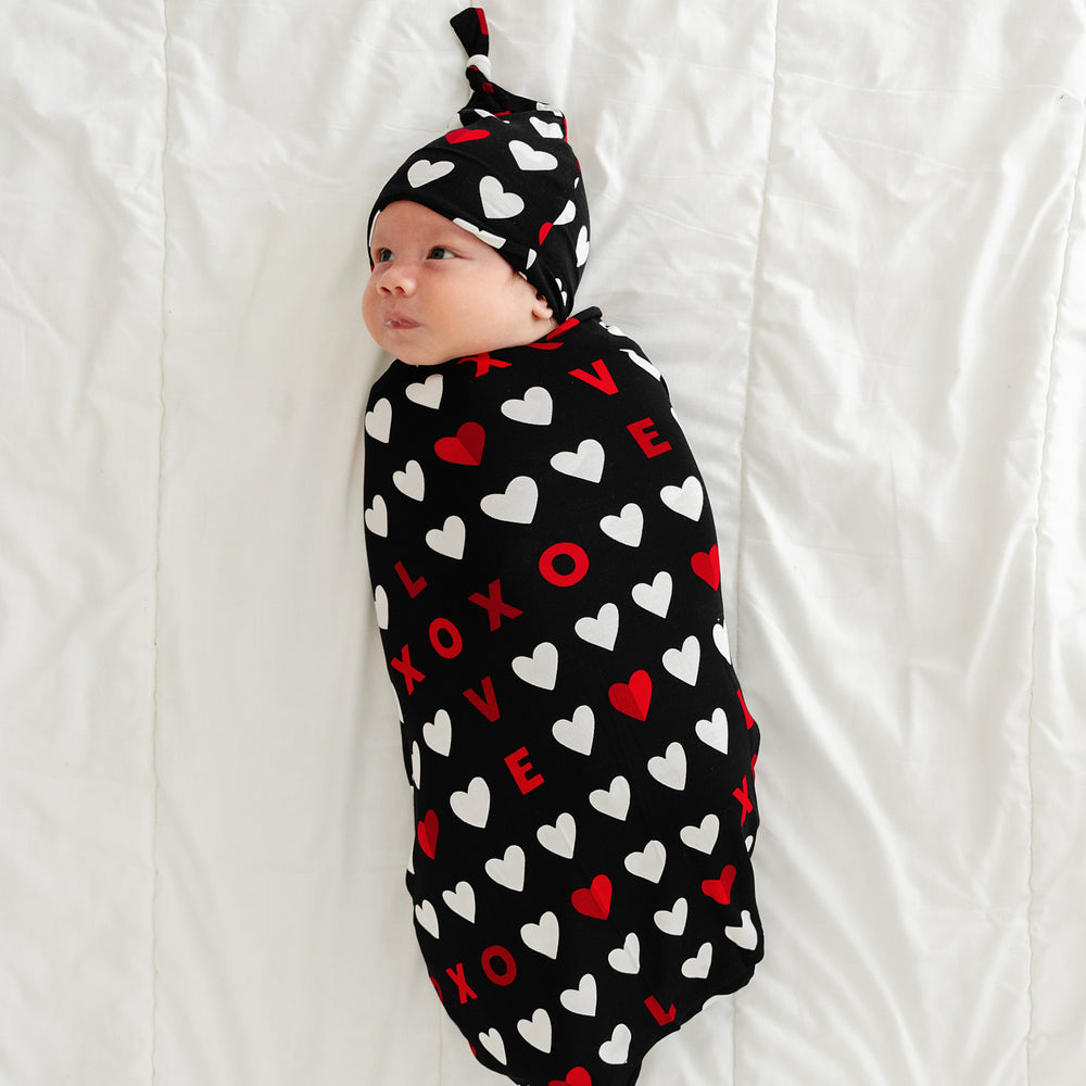 Click to see full screen - Child laying on a bed swaddled in a Black XOXO swaddle and hat set