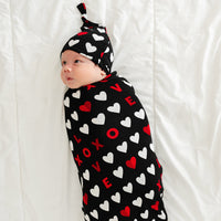 Alternate image of a child laying on a bed swaddled in a Black XOXO swaddle and hat set