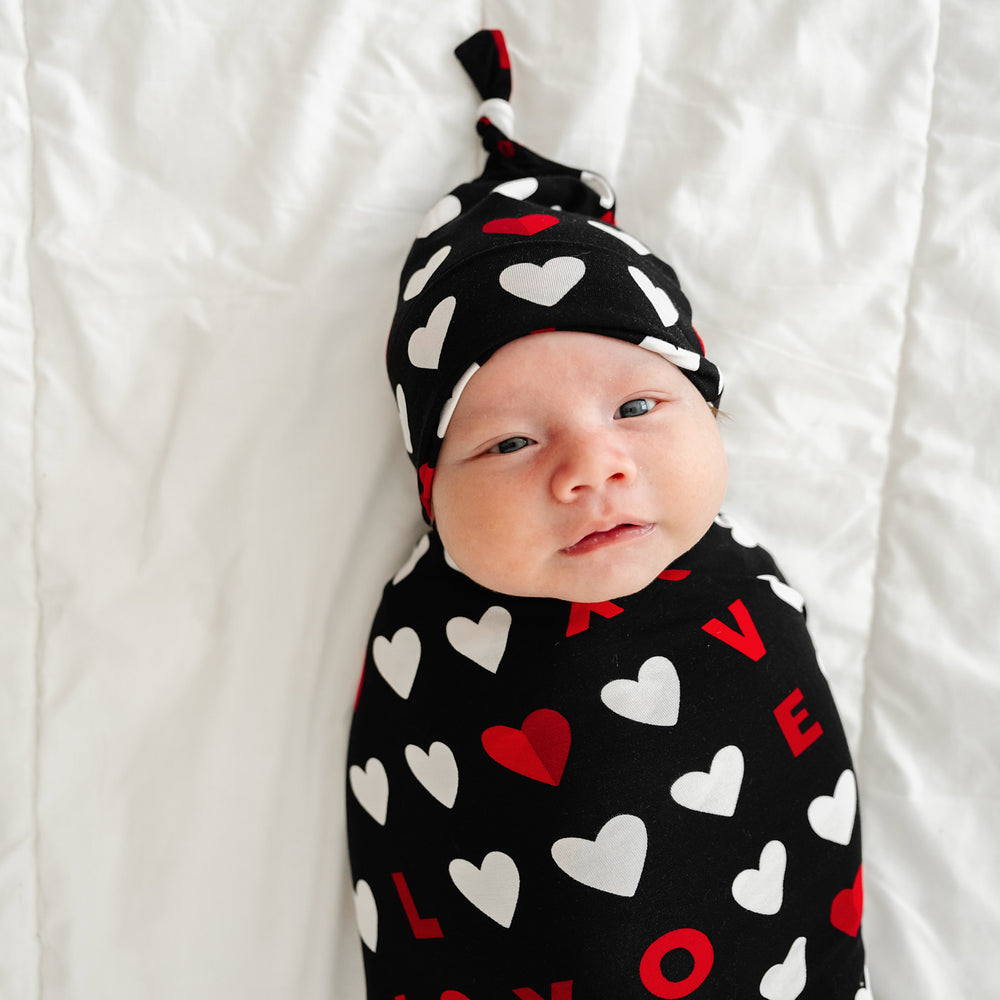 Click to see full screen - Close up image of a child laying on a bed swaddled in a Black XOXO swaddle and hat set