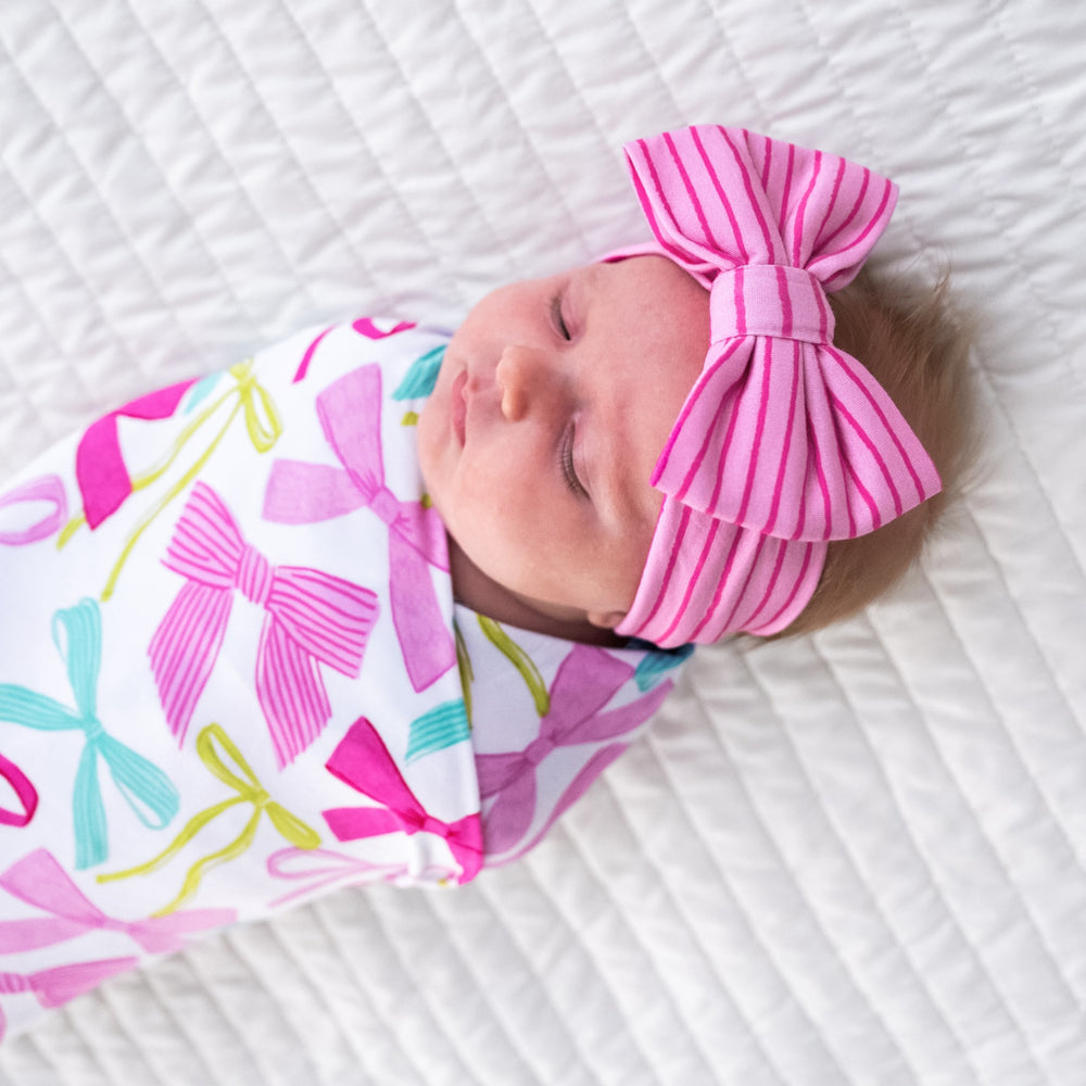 Close up image of a child swaddled on a bed in a Ribbons and Bows swaddle and luxe bow headband set