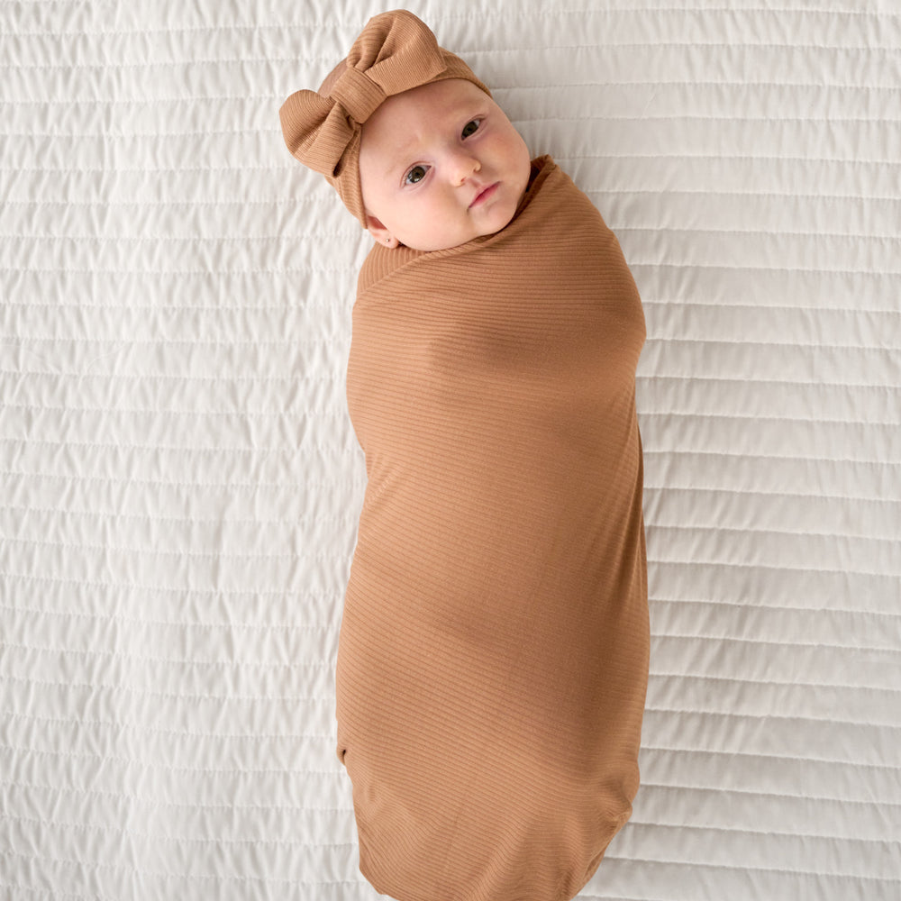 Top view image of baby laying down while wrapped in the Caramel Ribbed Swaddle & Luxe Bow Headband Set