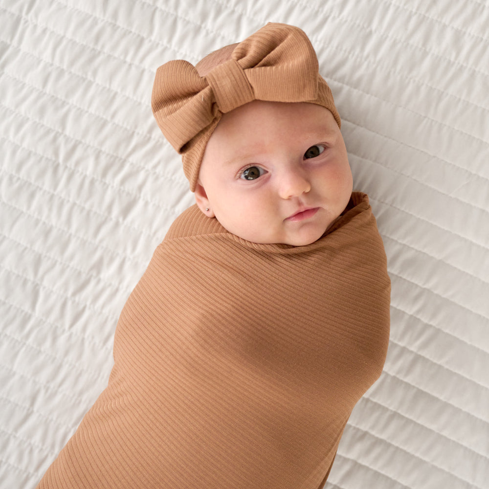 Close up image of baby laying down wearing the bow headband within the Caramel Ribbed Swaddle & Luxe Bow Headband Set