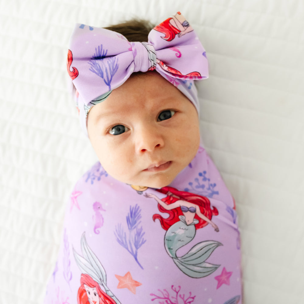 Close up image of a child swaddled in a Disney Part of Her World swaddle and luxe bow headband set