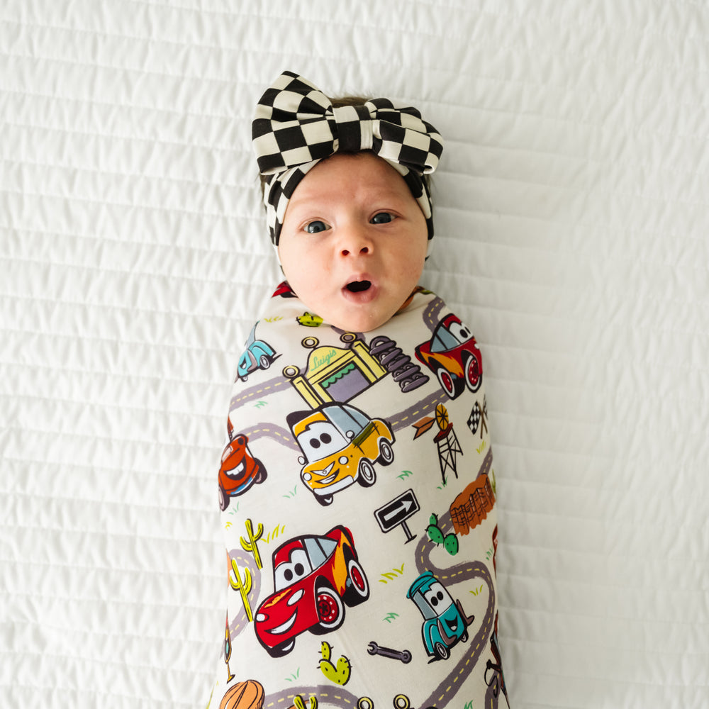Close up image of a child laying on a bed swaddled in a Radiator Springs swaddle and luxe bow set