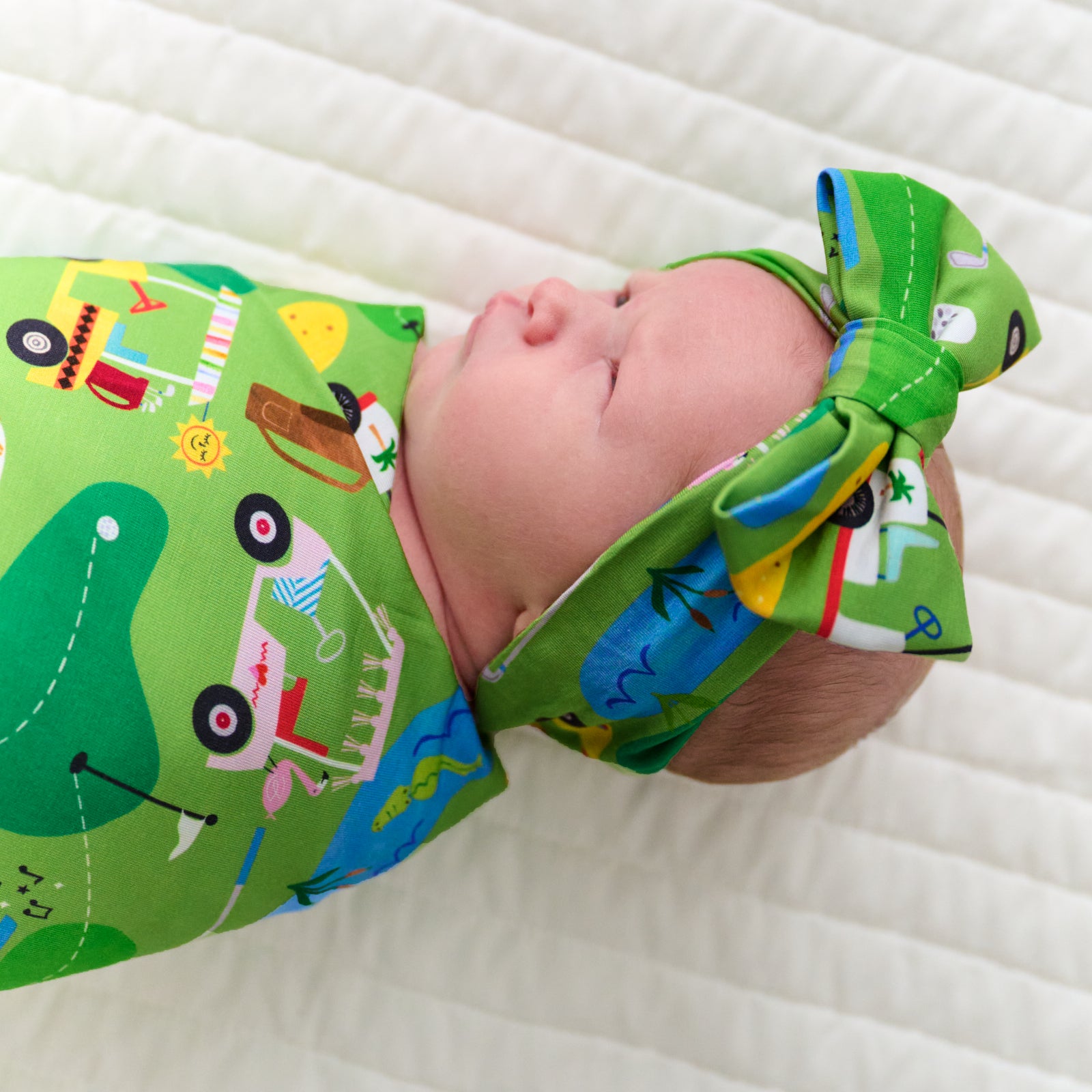 Close up image of an infant swaddled in a Fairway Fun swaddle and luxe bow headband set