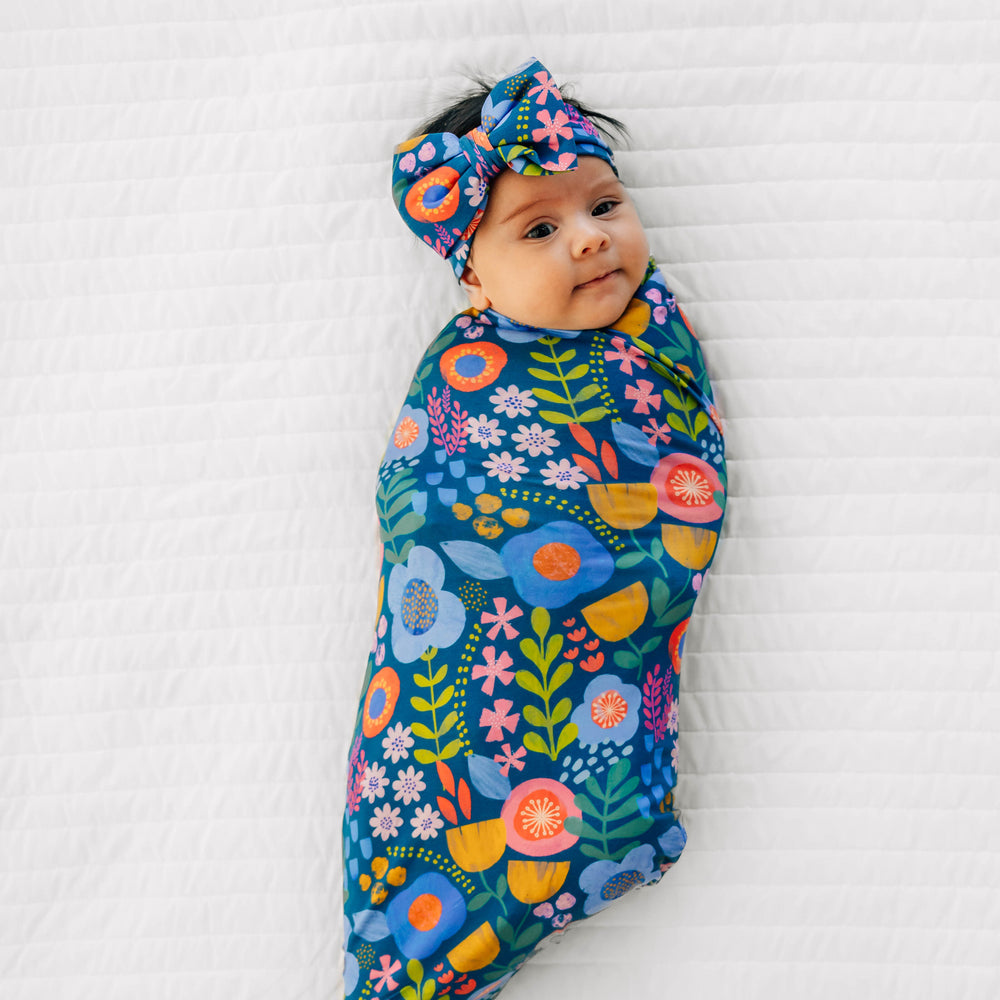 Top view image of baby laying with the Folk Floral Swaddle & Luxe Bow Headband Set