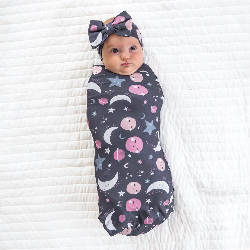 Child laying on a bed wearing Pink to the Moon and Back swaddle and luxe bow headband
