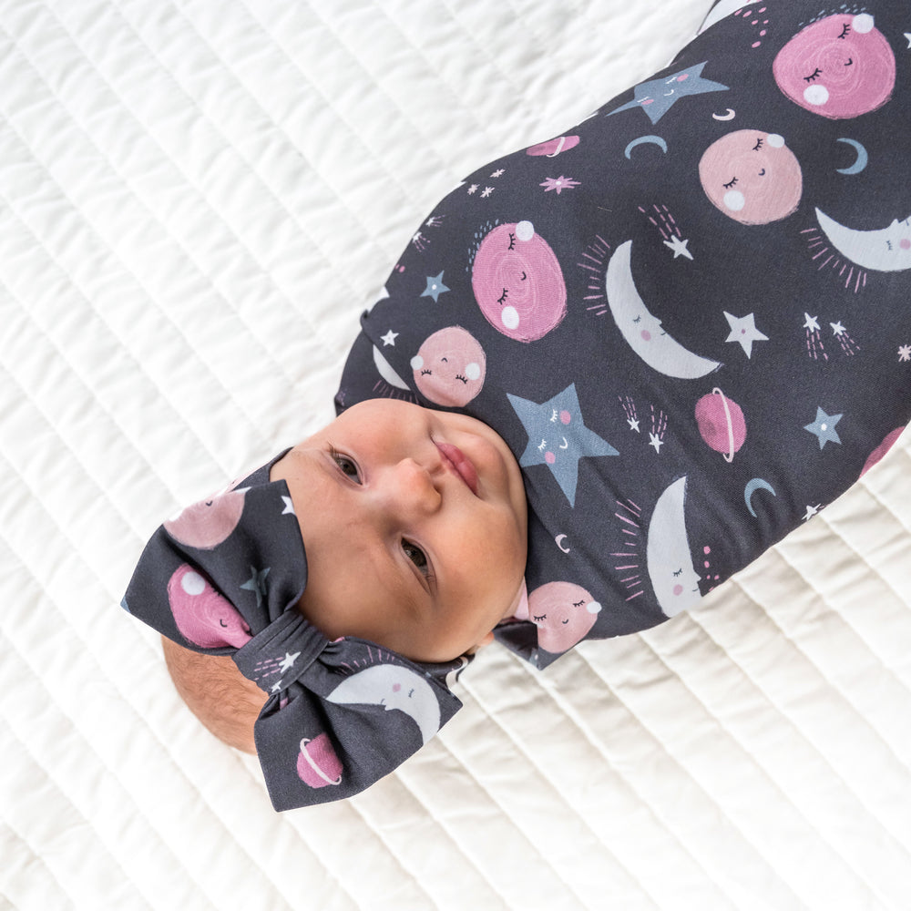 Close up image of a child laying on a bed wearing Pink to the Moon and Back swaddle and luxe bow headband
