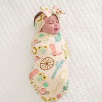 Child on a bed swaddled in a Pink Ready to Rodeo swaddle and luxe bow headband set