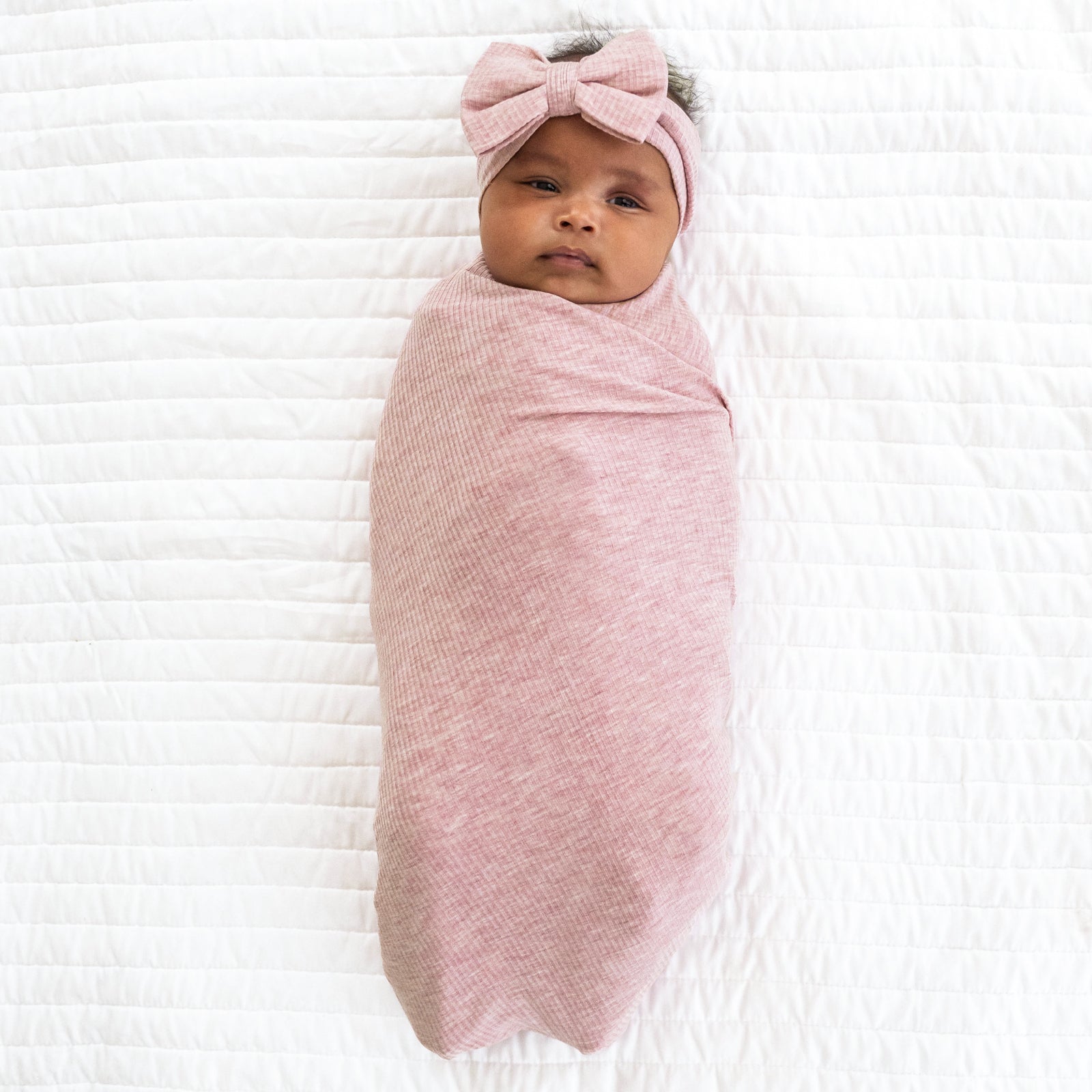 Child laying on a bed swaddled in a Heather Mauve swaddle and luxe bow headband set
