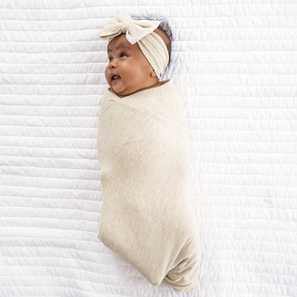 Image of a child swaddled in a Heather Oatmeal Ribbed Swaddle and Luxe Bow Headband Set