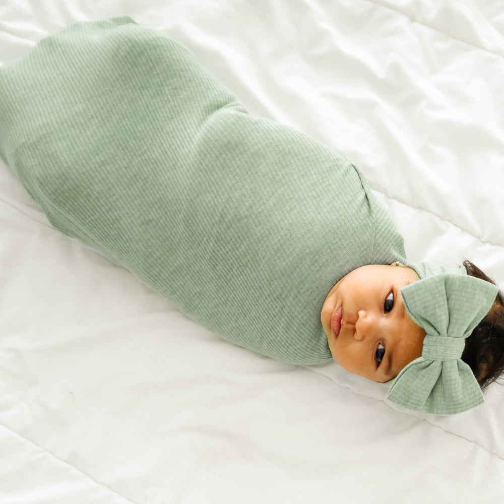 Click to see full screen - Alternate image of a child on a bed swaddled in a Heather Sage swaddle and Luxe bow Headband Set