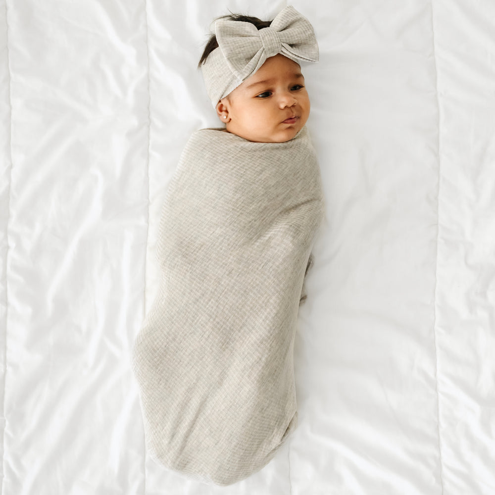 Click to see full screen - Child on a bed swaddled in a Heather Stone Ribbed swaddle and Luxe bow Headband Set