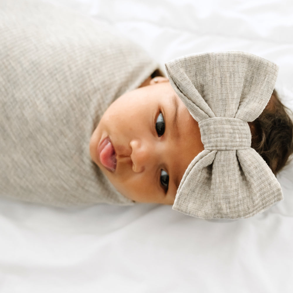 Close up image of a child on a bed swaddled in a Heather Stone Ribbed swaddle and Luxe bow Headband Set