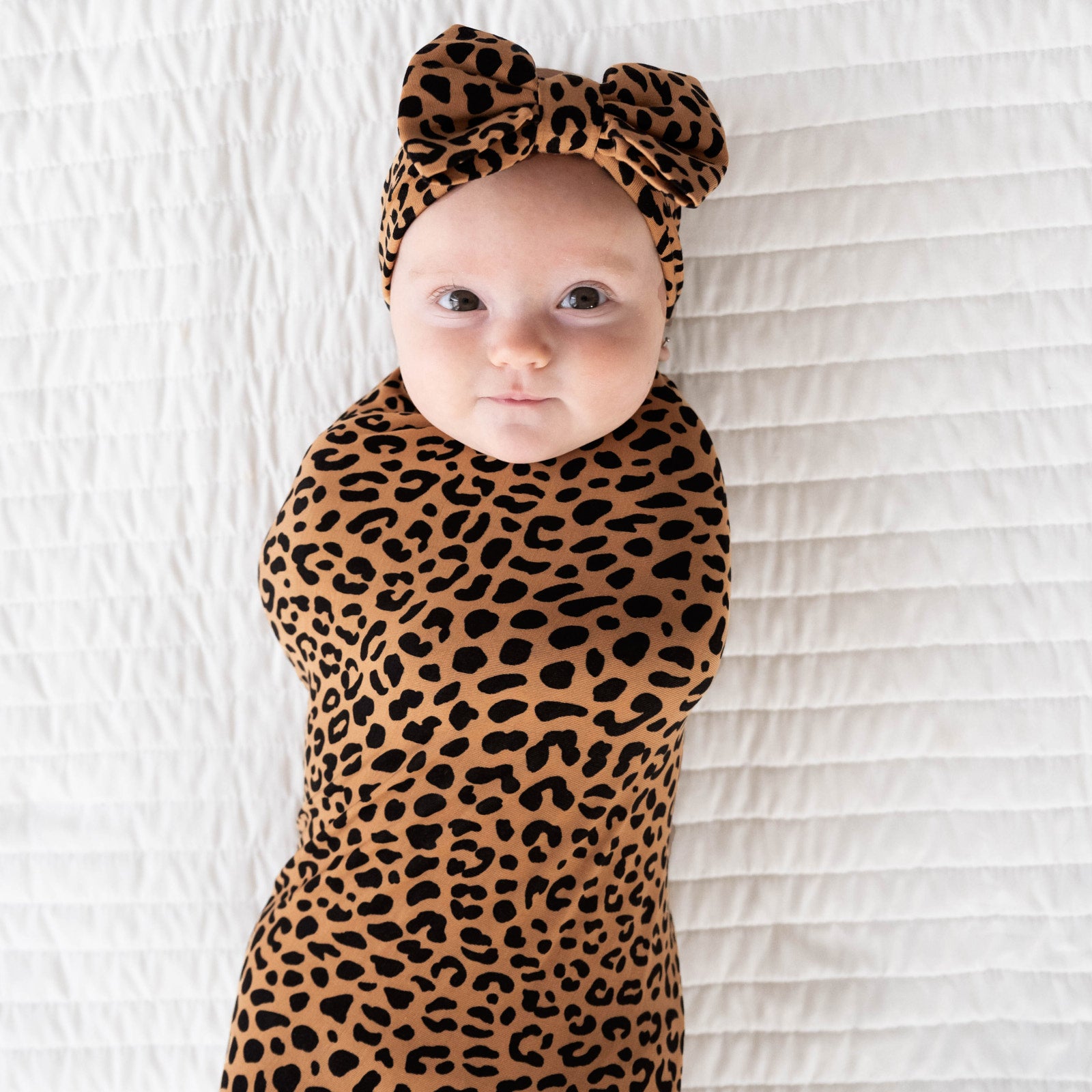 Alternative top view image of baby laying down while wearing the Classic Leopard Swaddle & Luxe Bow Headband Set