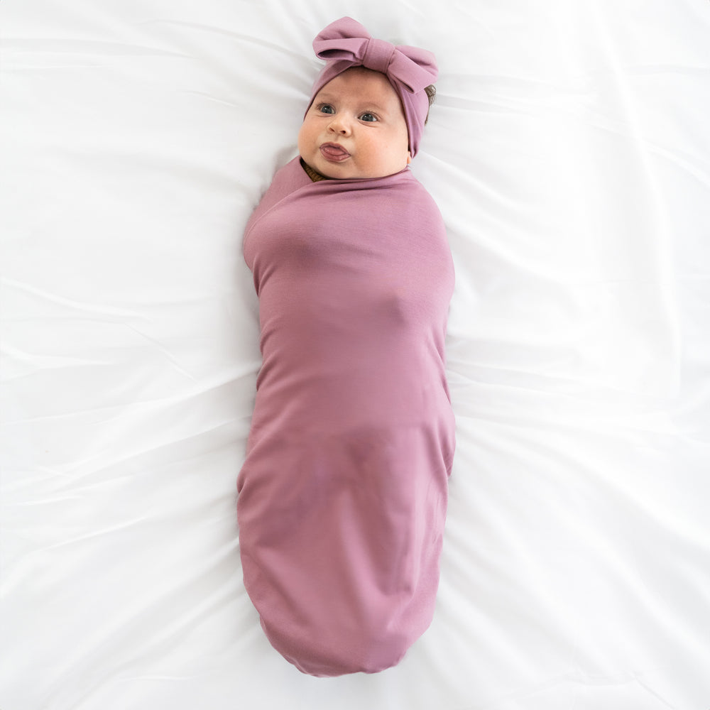 child swaddled on a bed in a Cozy dusty plum swaddle and luxe bow headband set