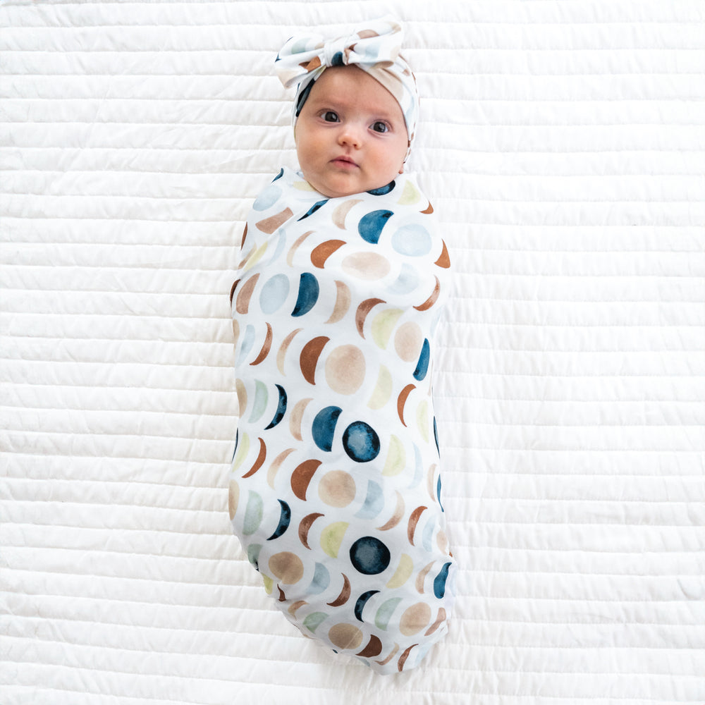 Alternate image of a child swaddled on a bed in a Luna Neutral swaddle and luxe bow headband set