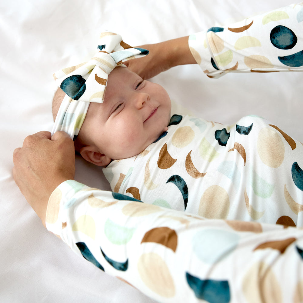 baby smiling wearing luna neutral swaddle with luxe bow headband as its being fixed by mom