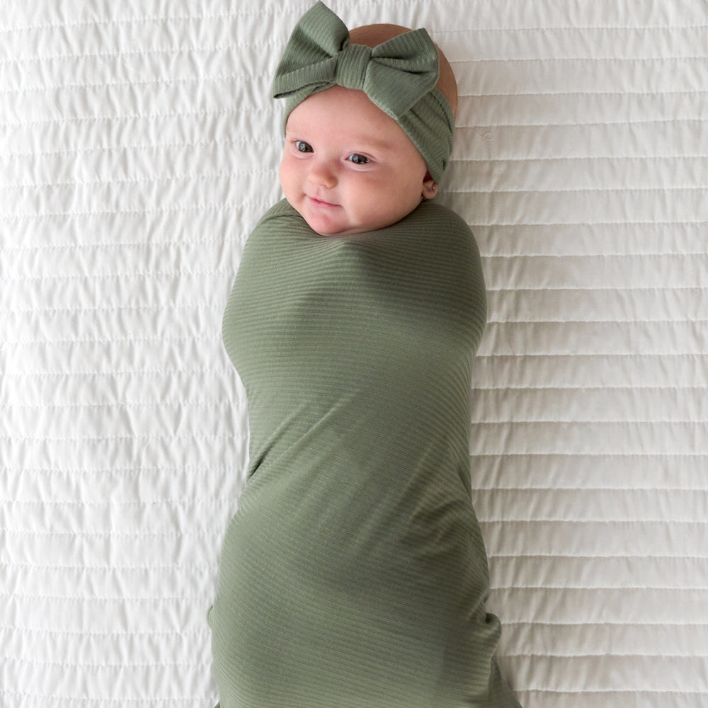 Top view image of baby laying down while wrapped in the Moss Ribbed Swaddle & Luxe Bow Headband Set