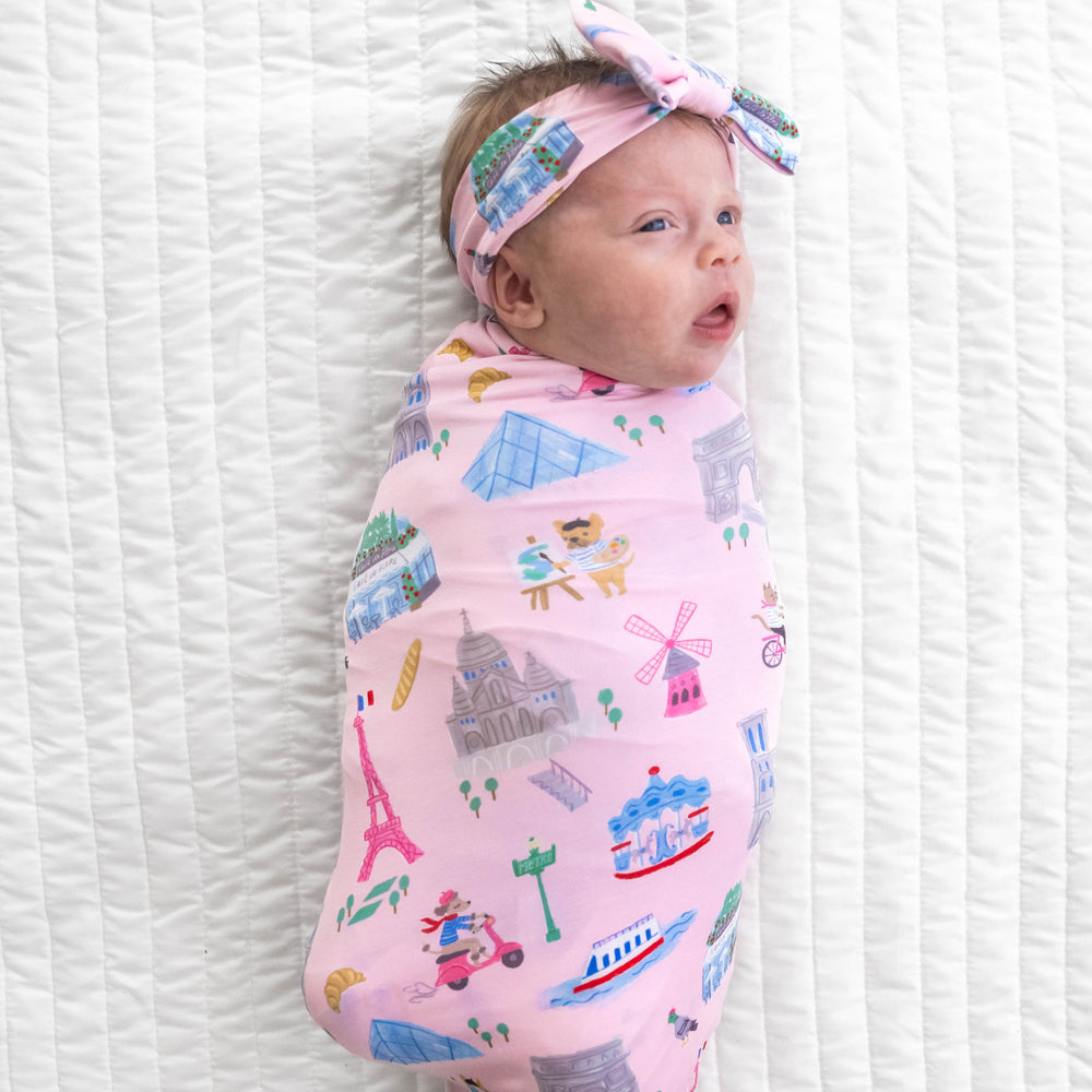 Top view image of baby laying down in the Pink Weekend in Paris Swaddle & Luxe Bow Headband Set