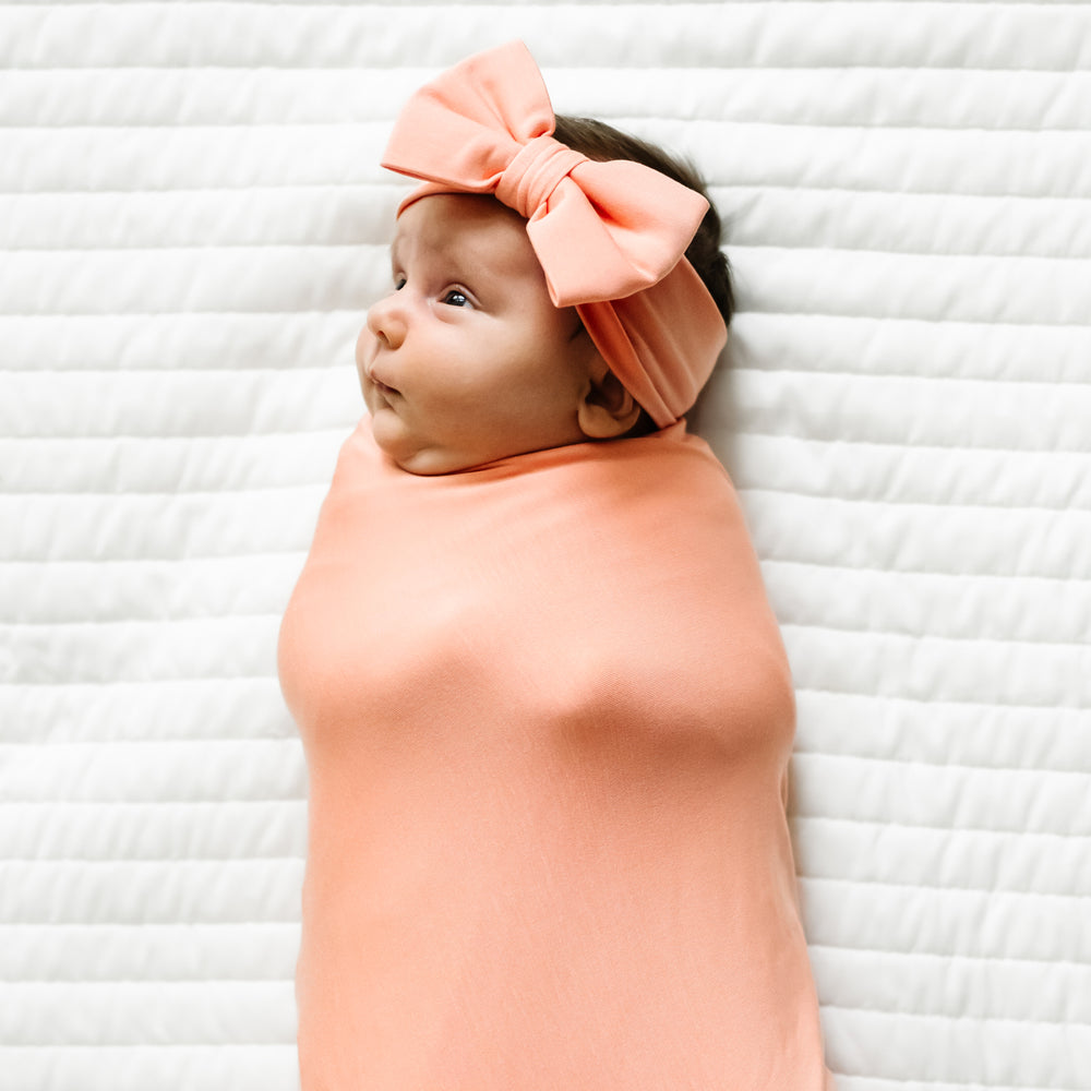 Child swaddled on a bed wearing a Peach swaddle and luxe bow headband set