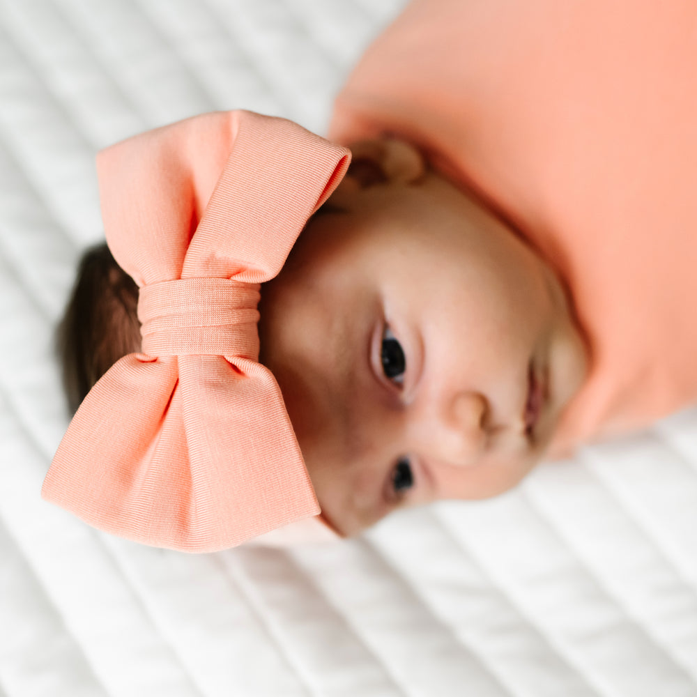 Close up image of a child swaddled on a bed wearing a Peach swaddle and luxe bow headband set