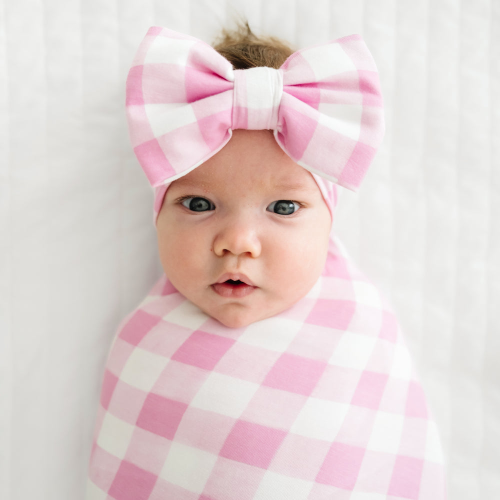 Click to see full screen - Close up image of a child swaddled in a Pink Gingham swaddle and luxe bow headband set
