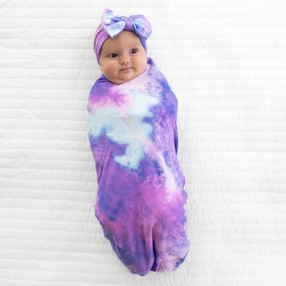 Child swaddled on a bed in a Purple Watercolor swaddle and luxe bow headband set