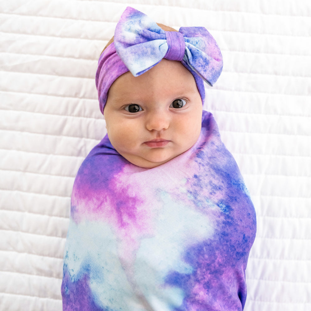 Close up image of a child swaddled on a bed in a Purple Watercolor swaddle and luxe bow headband set