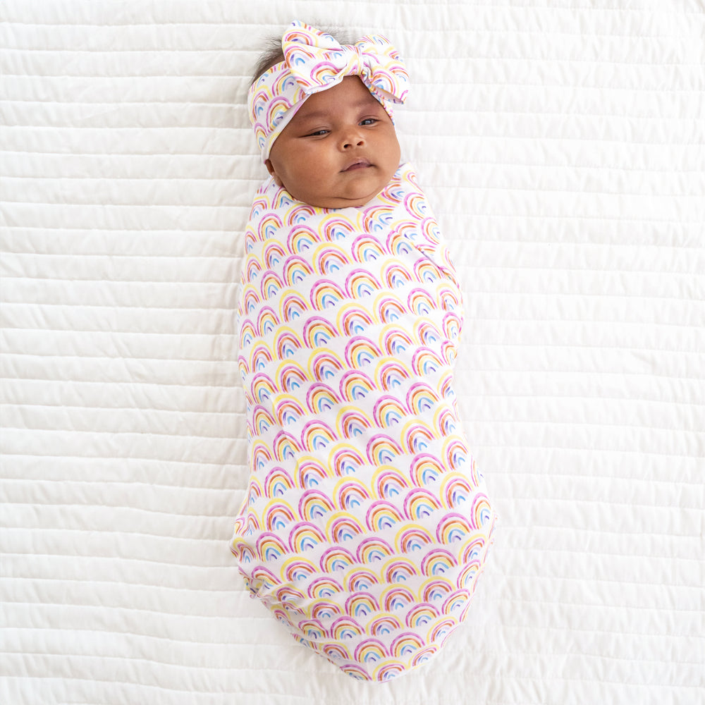 Child swaddled on a bed in a Pastel Rainbows swaddle and luxe bow headband set