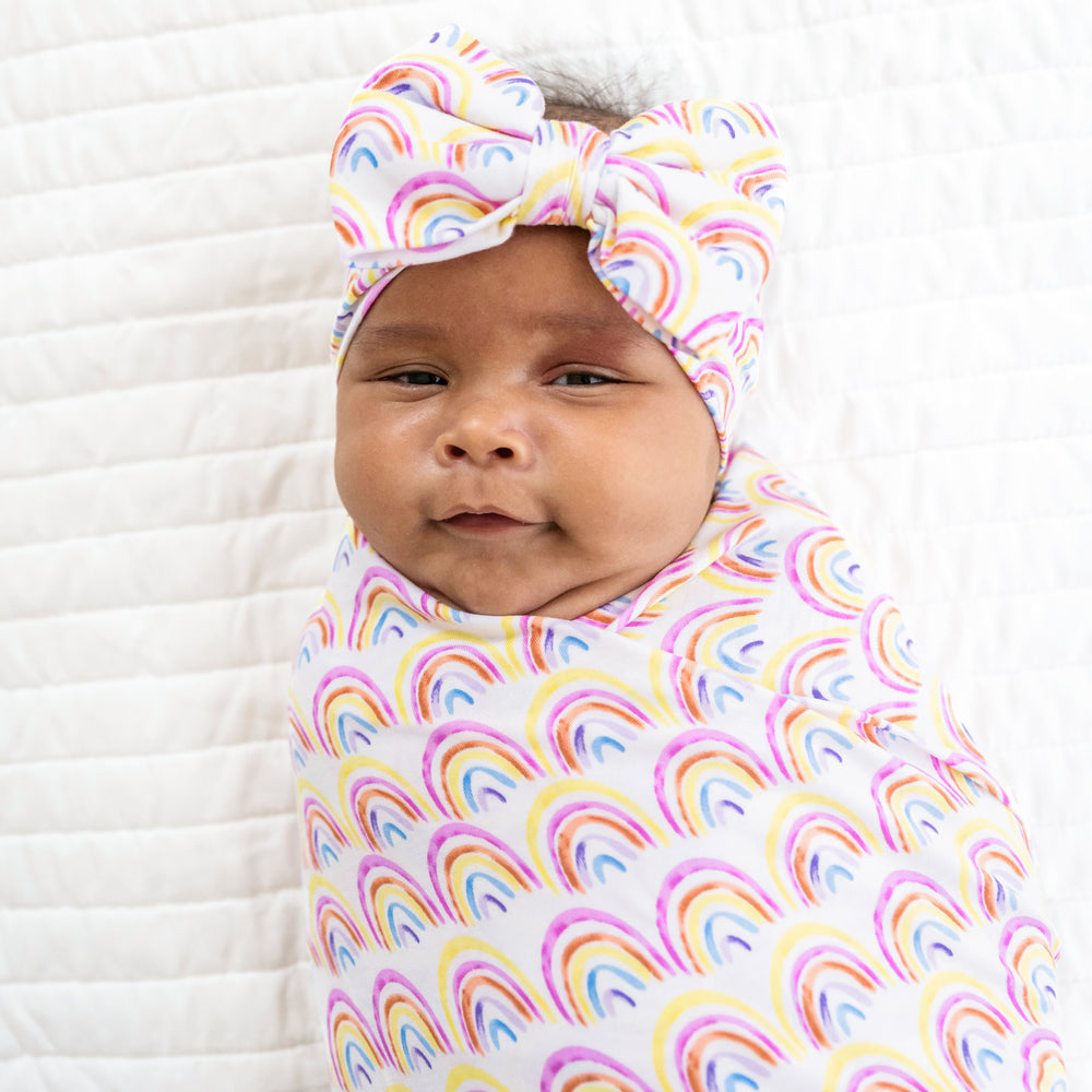 Close up image of a child swaddled on a bed in a Pastel Rainbows swaddle and luxe bow headband set