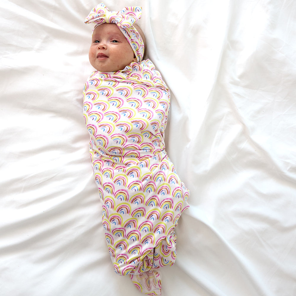 baby laying on bed wearing pastel rainbows swaddle set with headband