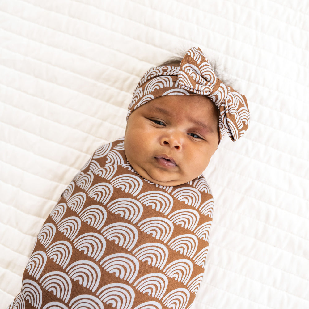 Close up image of a child swaddled on a bed in a Rust Rainbows swaddle and luxe bow headband set