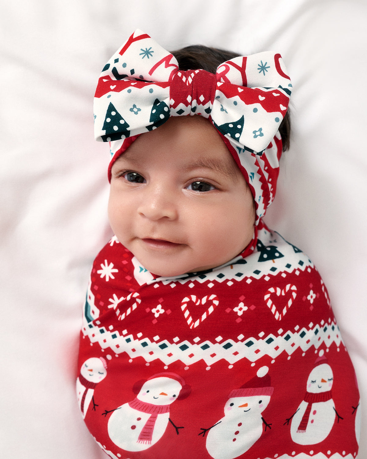 A baby wrapped in a festive red, white, and green holiday swaddle blanket, wearing a matching bow headband with a similar snowman and geometric pattern, lying on a white background.