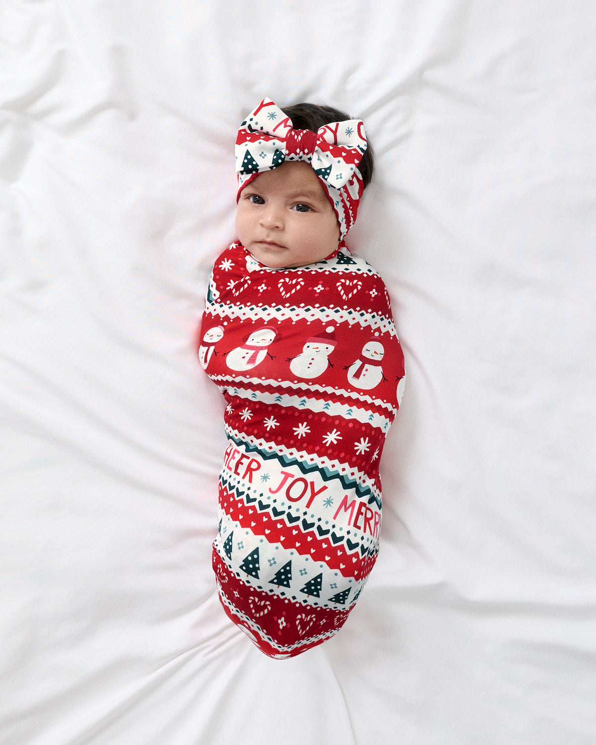 A baby lying on white bedding, swaddled in a festive red holiday wrap with snowman and Christmas tree patterns, and wearing a matching red bow headband.