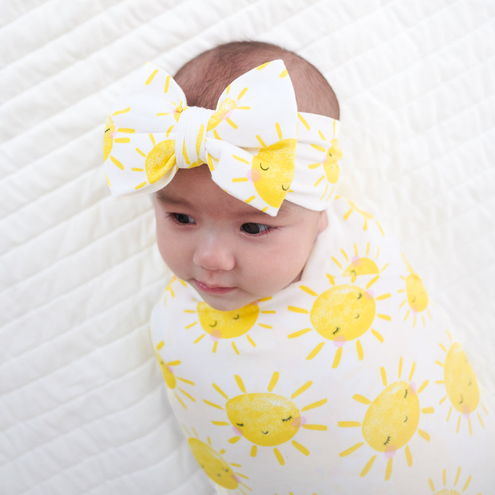 Close up image of a child laying on a bed swaddled in a Sunshine swaddle and luxe bow headband set
