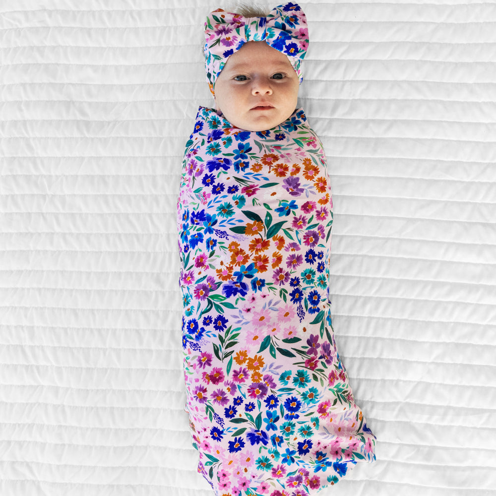 Image of a child swaddled on a bed in a Sweet Pea Floral swaddle and luxe bow headband